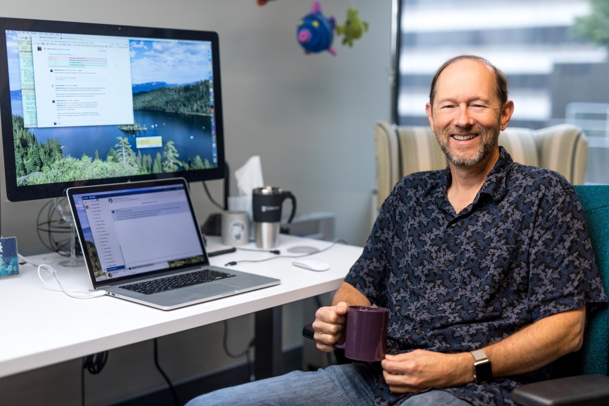Portrait of older male developer in shirt sleeve shirt, smiling and facing camera. He is sitting at a desk in an office setting, holding a coffee mug.