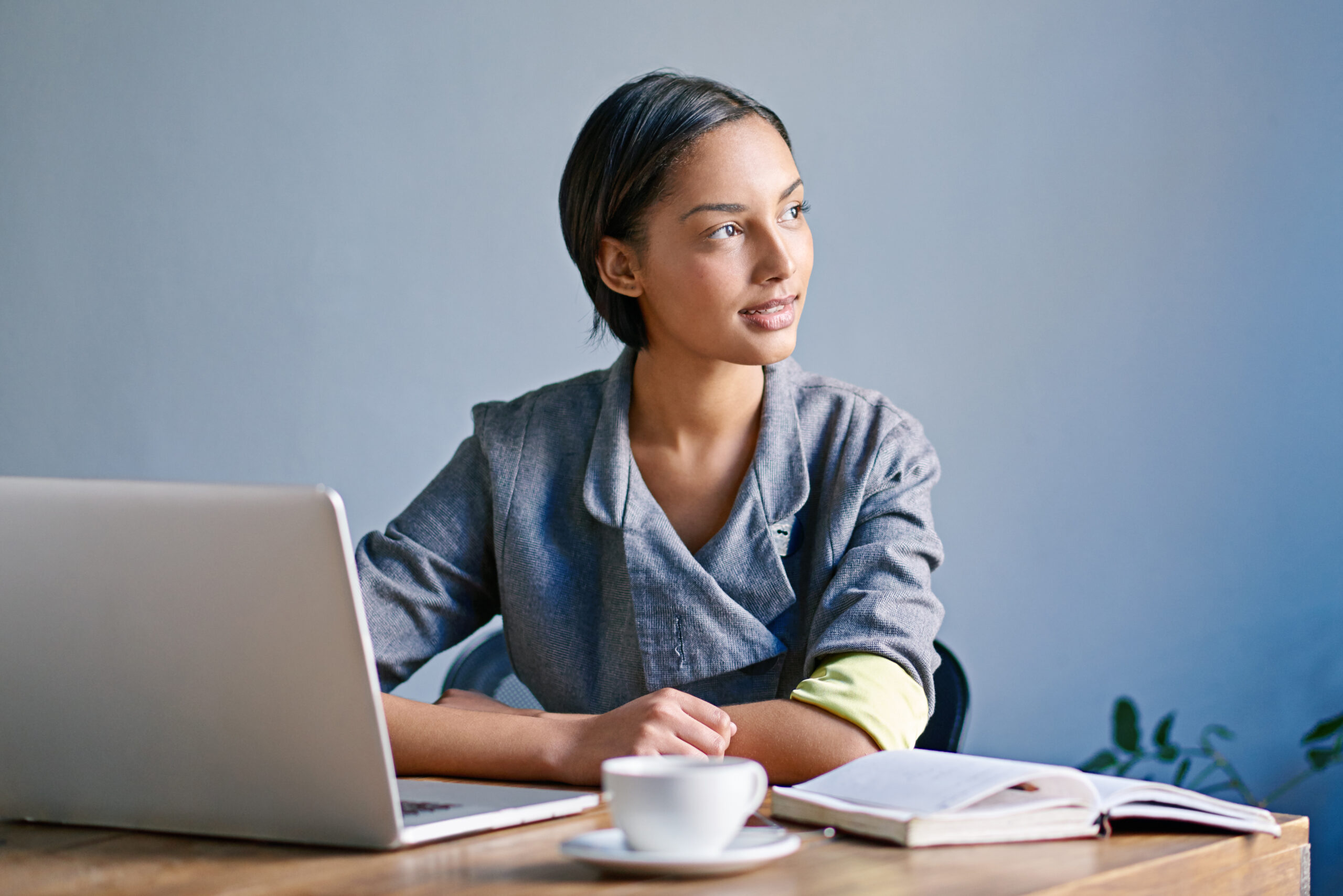 A woman is sitting at a wooden table with a laptop, an open book, and a cup of coffee. The background is plain and the person is wearing a grey jacket.
