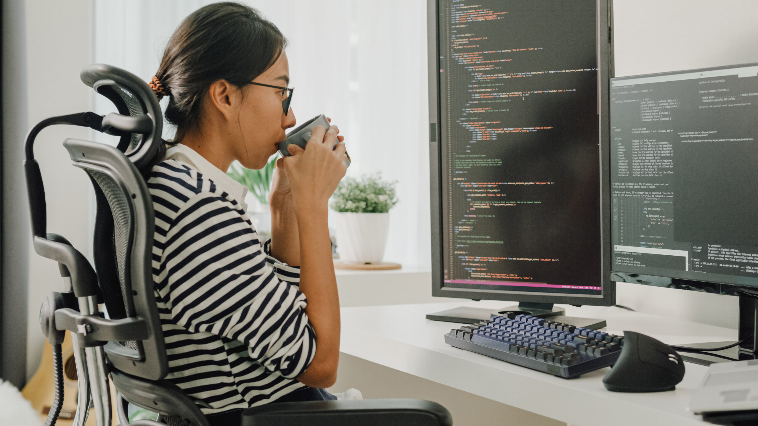 A software developer is using a computer to write code. She is sitting at desk with multiple monitors while drinking coffee.