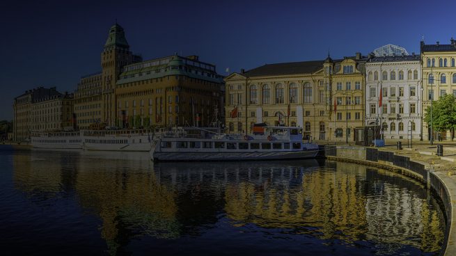 Warm summer sunlight illuminating the ferries on the harbour waterfront reflecting the iconic facades of the luxury hotels and townhouses that line the shore of Nybroviken harbour, Stockholm, Sweden's vibrant capital city.