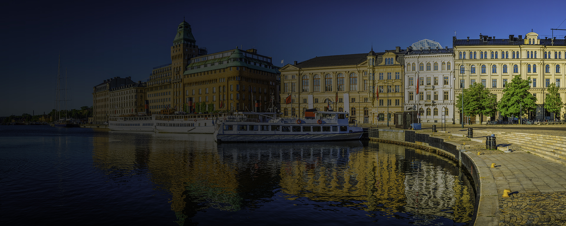 Warm summer sunlight illuminating the ferries on the harbour waterfront reflecting the iconic facades of the luxury hotels and townhouses that line the shore of Nybroviken harbour, Stockholm, Sweden's vibrant capital city.