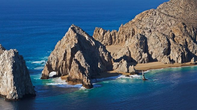 a rocky island in the middle of a body of water with Arch of Cabo San Lucas in the background