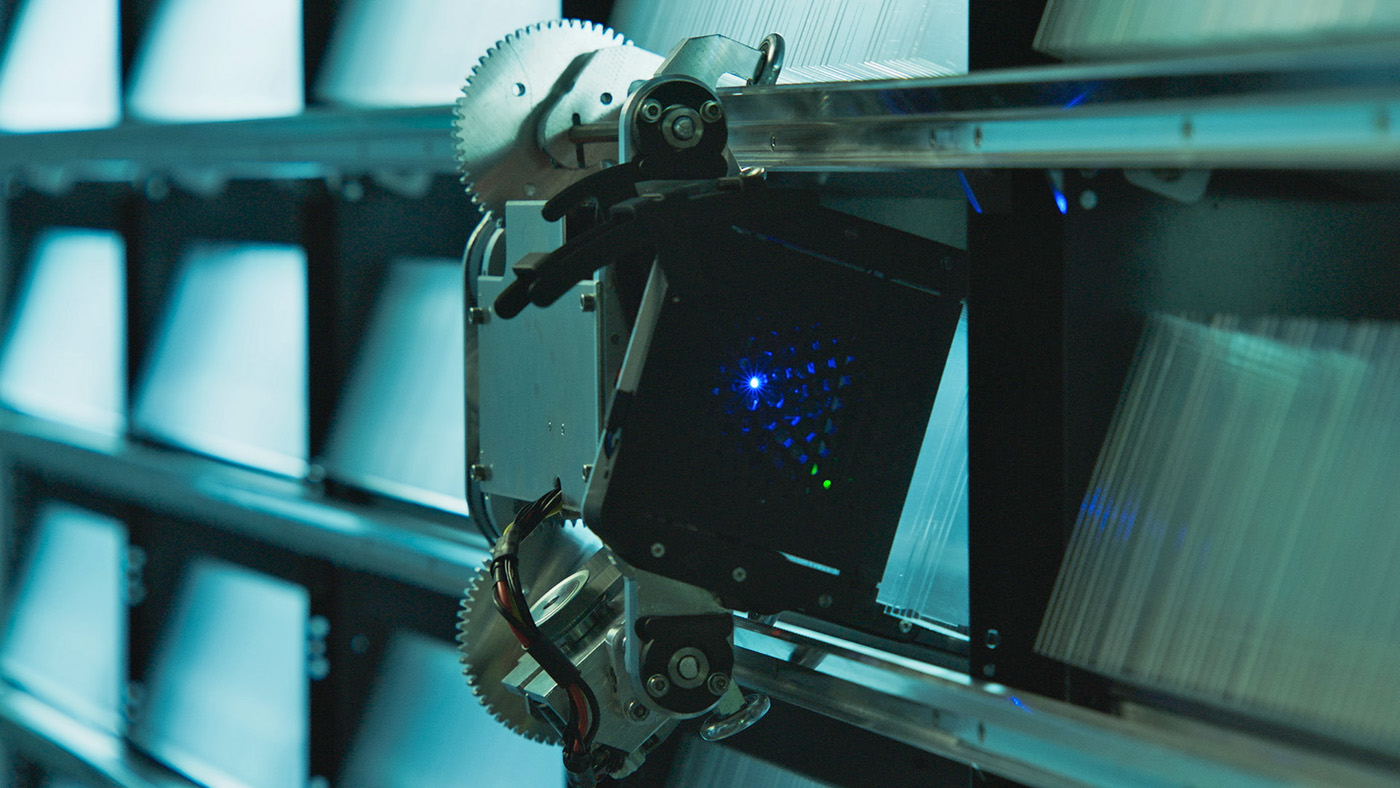 Picture of a RASCAL prototype mounted on a Silica library. The library is composed of a series of connected storage racks that hold glass media. The storage panel's front has parallel rails mounted horizontally to allow the robot to move vertically and horizontally. RASCAL uses a pair of opposing wheels to latch onto these rails. 