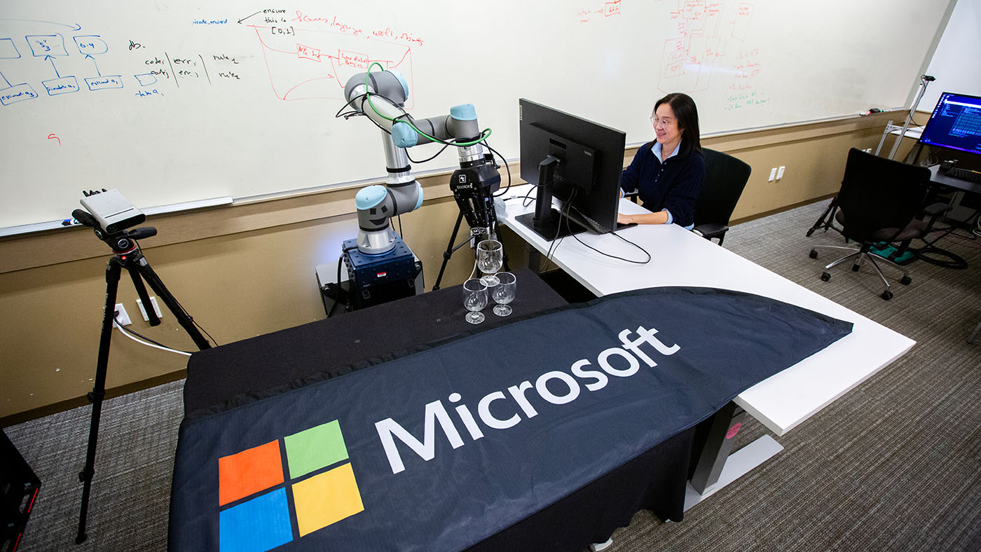 Photo of a woman sitting at a Microsoft table in front of a computer screen while demonstrating a robotic arm picking up a glass