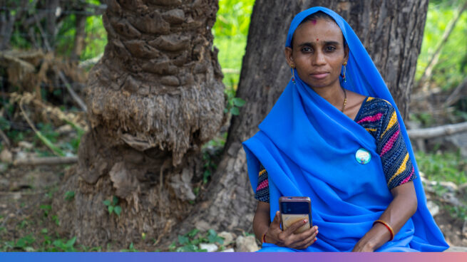 Lady in blue sari, holding a smartphone, looking at the camera. Seated outside in front of a couple of trees.