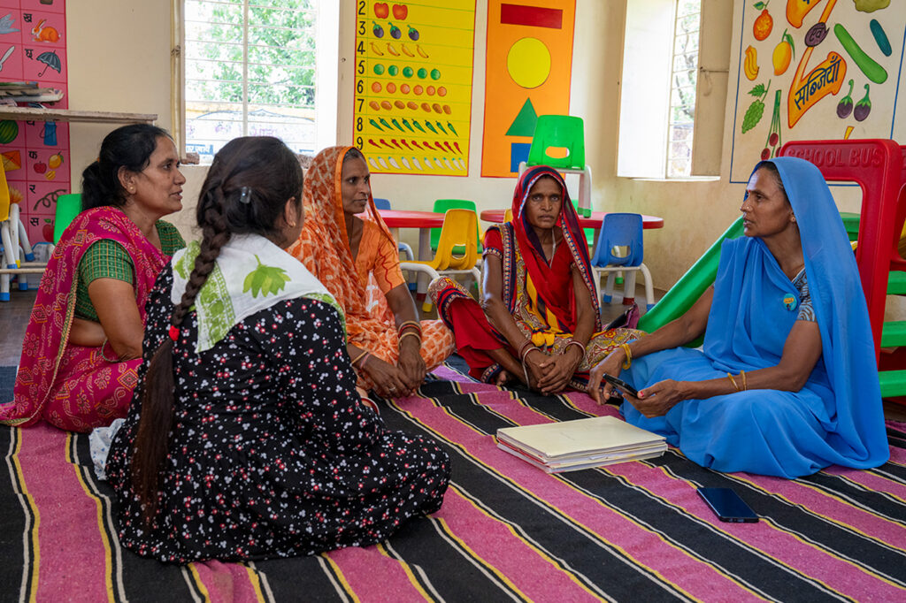 Five ladies in colorful saris seated on a rug at a classroom talking. The lady on the far right, wearing a blue sari, is holding a smartphone, and has a stack of papers at in front of her.​