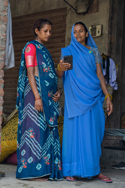 One lady in blue sari talking to ASHA standing outside of a brick house, one of them holding a smartphone.​