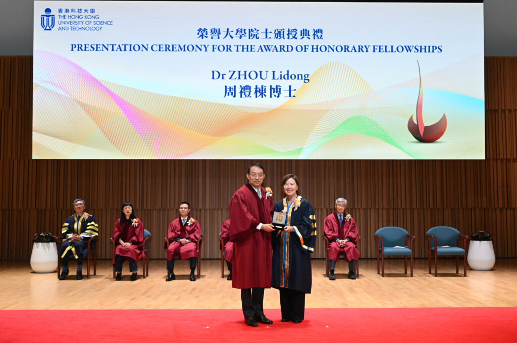 A presentation ceremony at The Hong Kong University of Science and Technology for the award of honorary fellowships. Lidong Zhou receives an award from HKUST President Prof. Nancy IP on stage, both dressed in academic regalia. A large screen above displays the event title and Lidong’s name in English and Chinese, with colorful abstract graphics.
