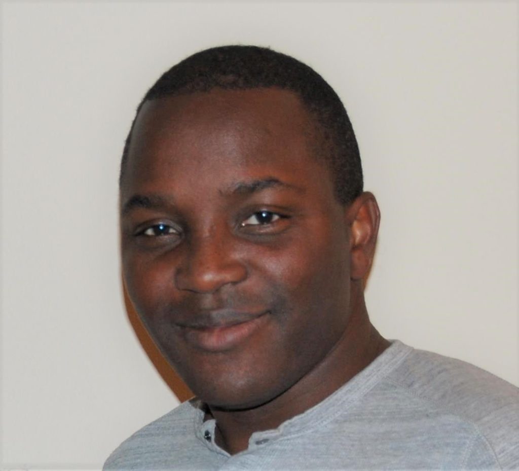 A profile headshot of Joe Dadzie, wearing a grey shirt against a cream-colored wall. 
