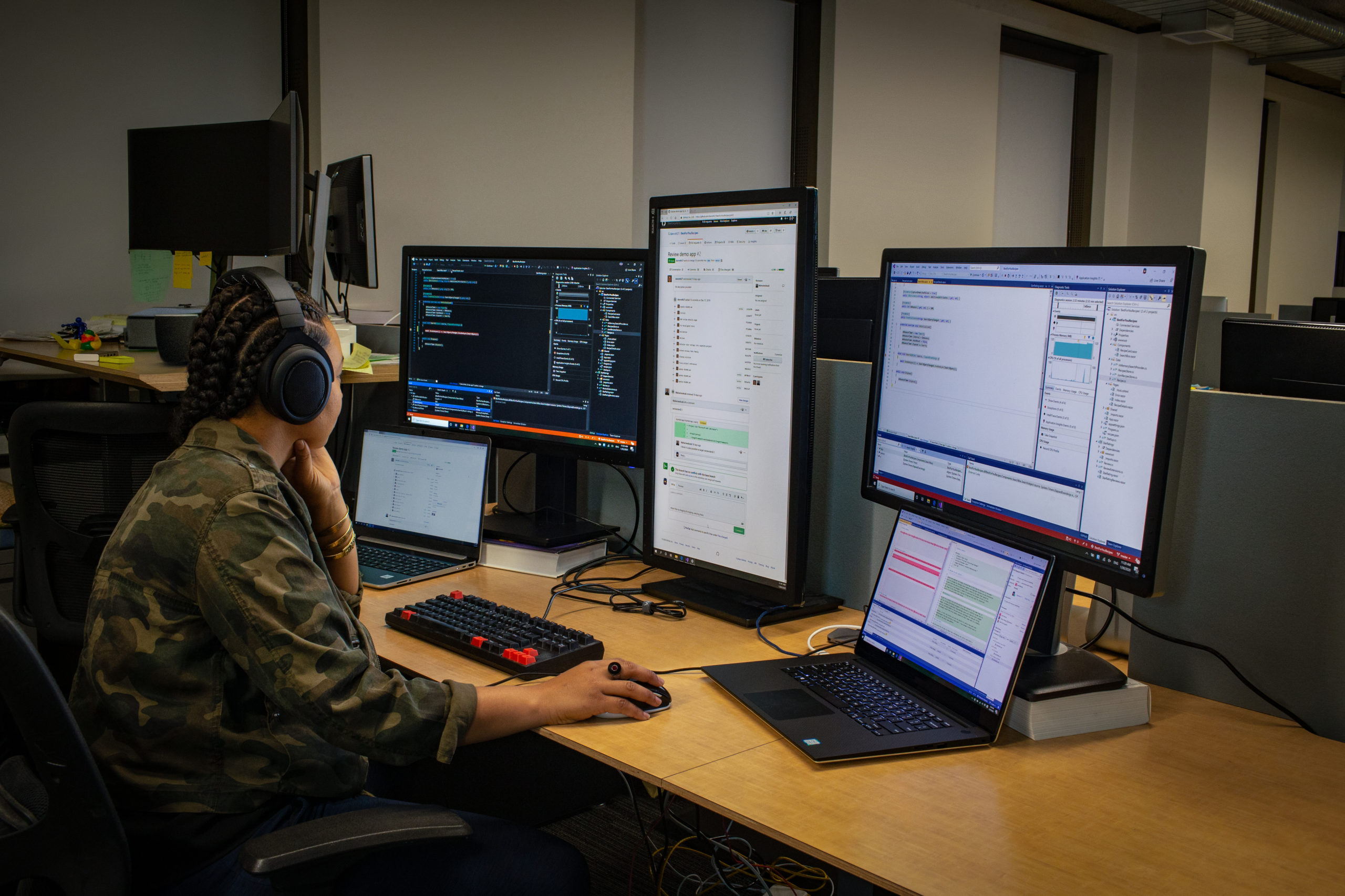 lack female developer wearing headphones; coding at her PC workspace in an enterprise office, using Visual Studio on a multi-monitor set up.