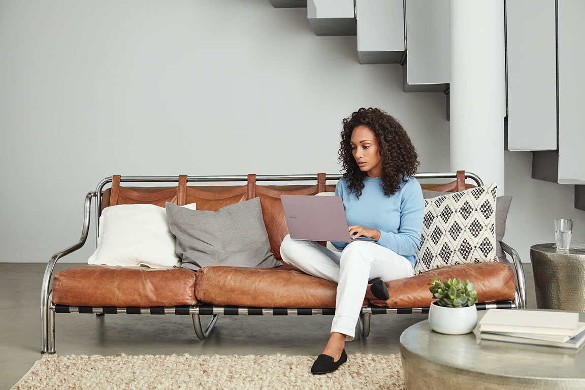 Adult inside home working, sitting on a brown couch, looking at a computer.