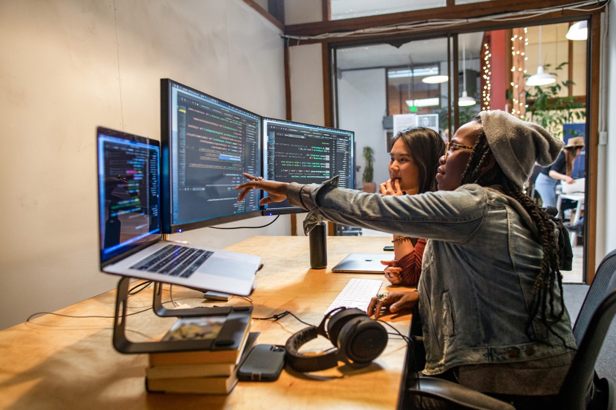 Two female developers working and collaborating in an enterprise office. They are using Visual Studio on a MacBook Pro with a multi-monitor set up.