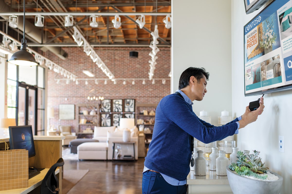 Male employee in commercial retail store plugging Xogo IoT device into wall-mounted monitor. The screen shows product promo ads for home furnishings.