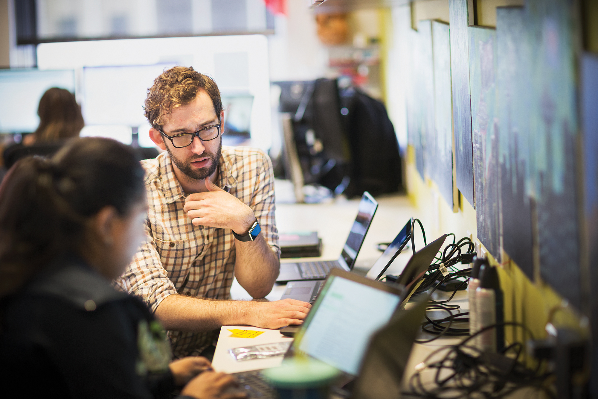 A man standing and looking at a colleague who is typing on a laptop.
