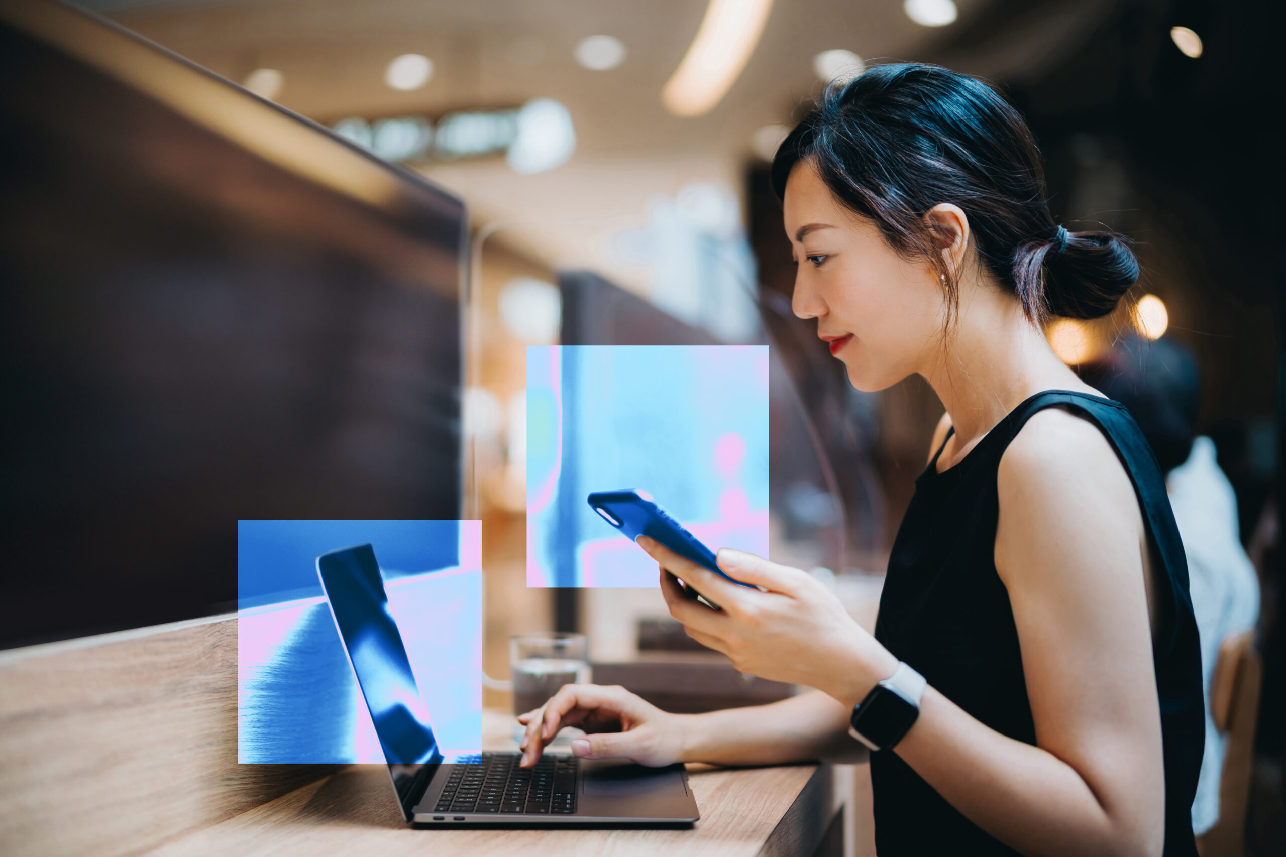 A woman looks at her computer screen and holds a phone in her left hand