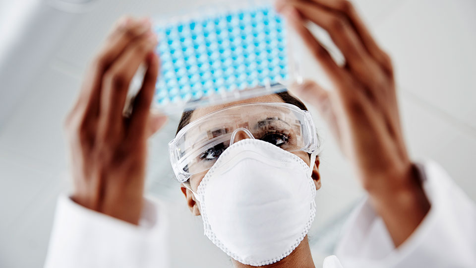 A medical professional in safety gear examines a rack of samples