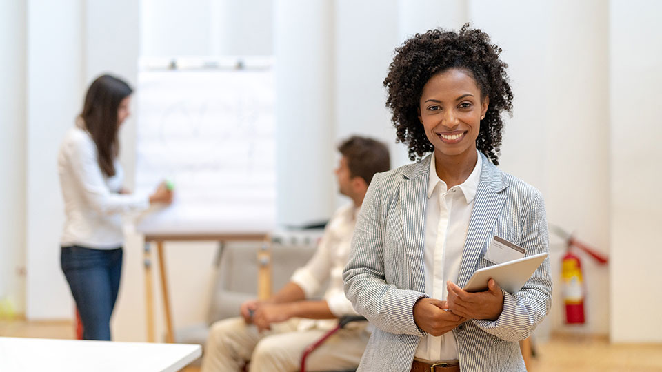 A smiling woman holding a tablet stands in front of a pair of coworkers in a discussion