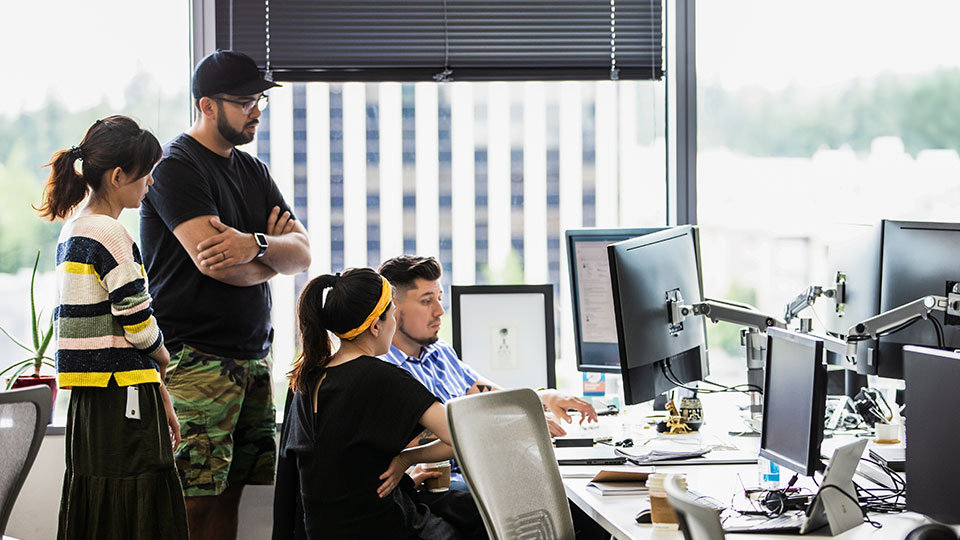 A group of coworkers in a casual office environment collaborate at a computer workstation