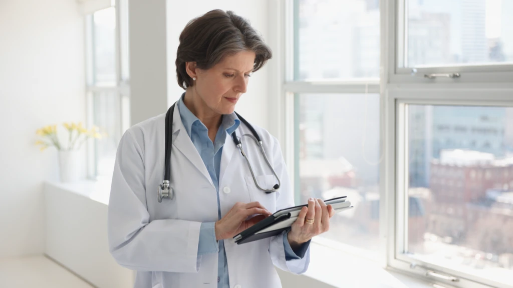 A doctor in a white lab coat uses a tablet while standing beside large windows inside a bright healthcare facility.