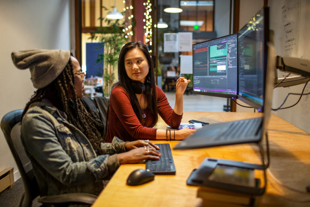 Two people collaborating at a shared desk with large monitors displaying code in a modern office.