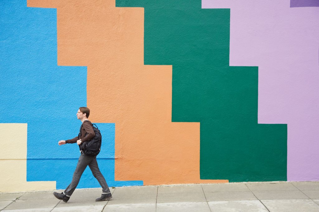 Photographie d’un homme marchant le long d’un trottoir devant une mosaïque murale colorée.