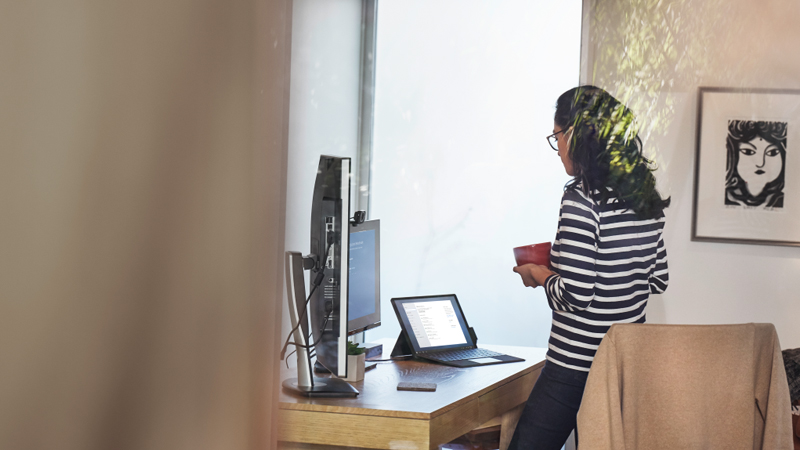 Femme travaillant depuis son bureau à domicile sur un appareil Surface Pro, debout à son bureau avec une tasse dans les mains