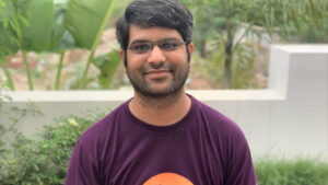 Kumar poses outside wearing a purple t-shirt, smiling towards the camera.