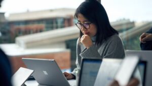 Woman with glasses sitting at a community table in common area reading on a Surface laptop.