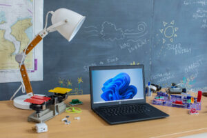 An open laptop sits on a table in a school classroom surrounded by science supplies with a blackboard in the background.