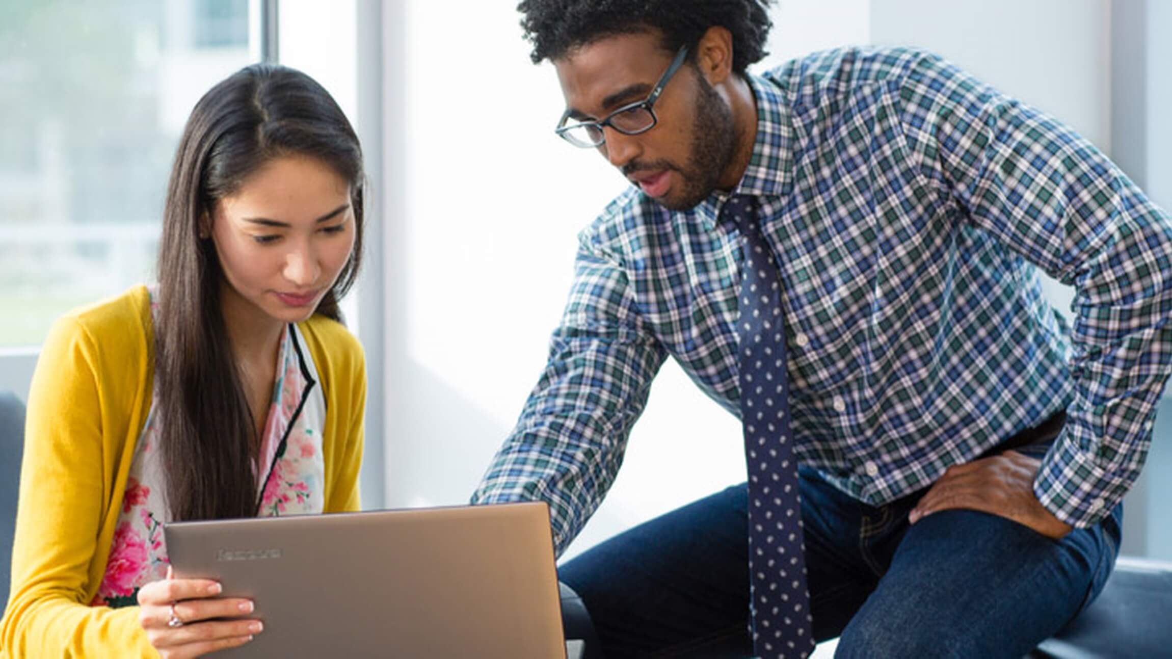Two teachers talk over a lesson plan while sitting at a desk looking at a PC.