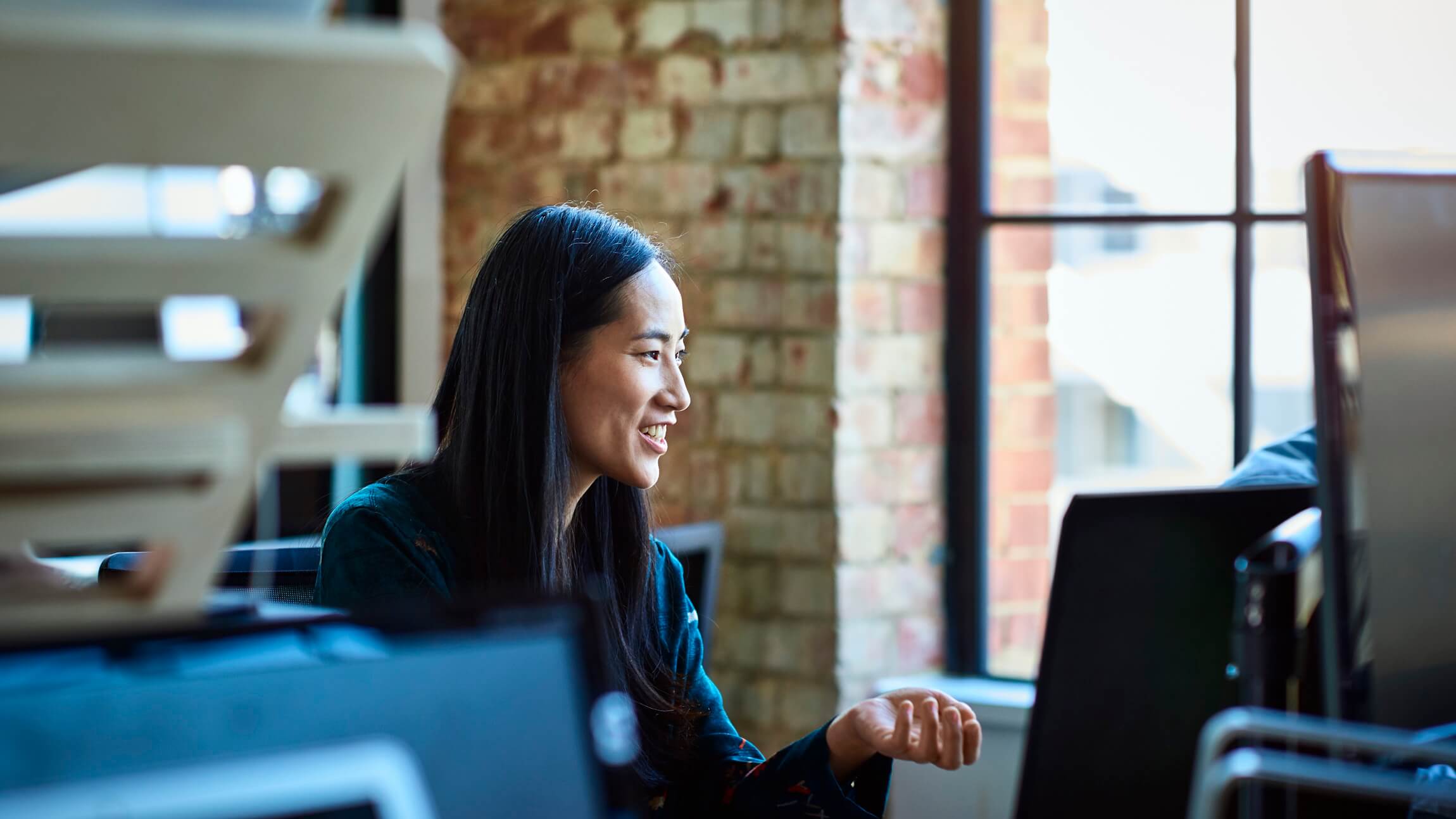 A woman sits at her workstation in a Microsoft office and chats with someone off camera nearby.