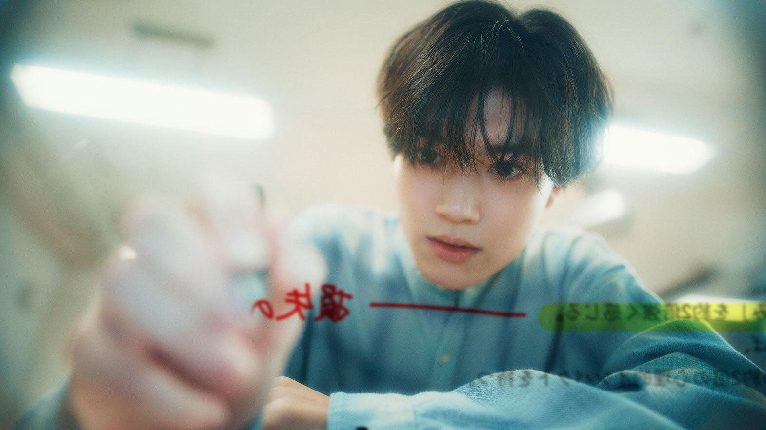 透明なガラス面やスクリーンに向かってペンで書き込みをしている、真剣な表情の若い男性。視聴者側からは、赤と緑の手書き文字が反転して見えている。