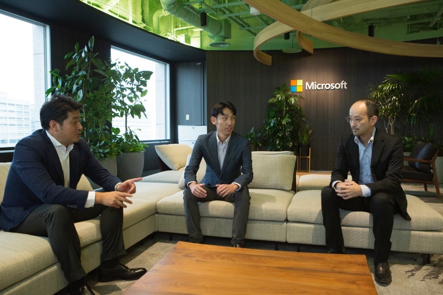 Microsoftロゴをバックにプロジェクトについて語る日本マイクロソフト社員とクライアント