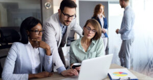 財務グラフが表示されたノート PC とタブレットをデスクで座って見ている 2 人の女性の財務専門家と、後ろから身を乗り出してノート PC 上で何かを操作している 1 人の男性の財務専門家。