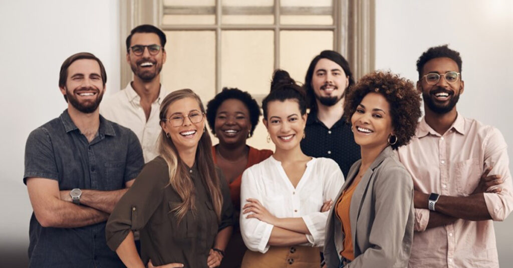 カジュアルな装いで会議室の端に集合して微笑む、民族的なルーツも年齢も異なる 8 人のプロフェッショナル。