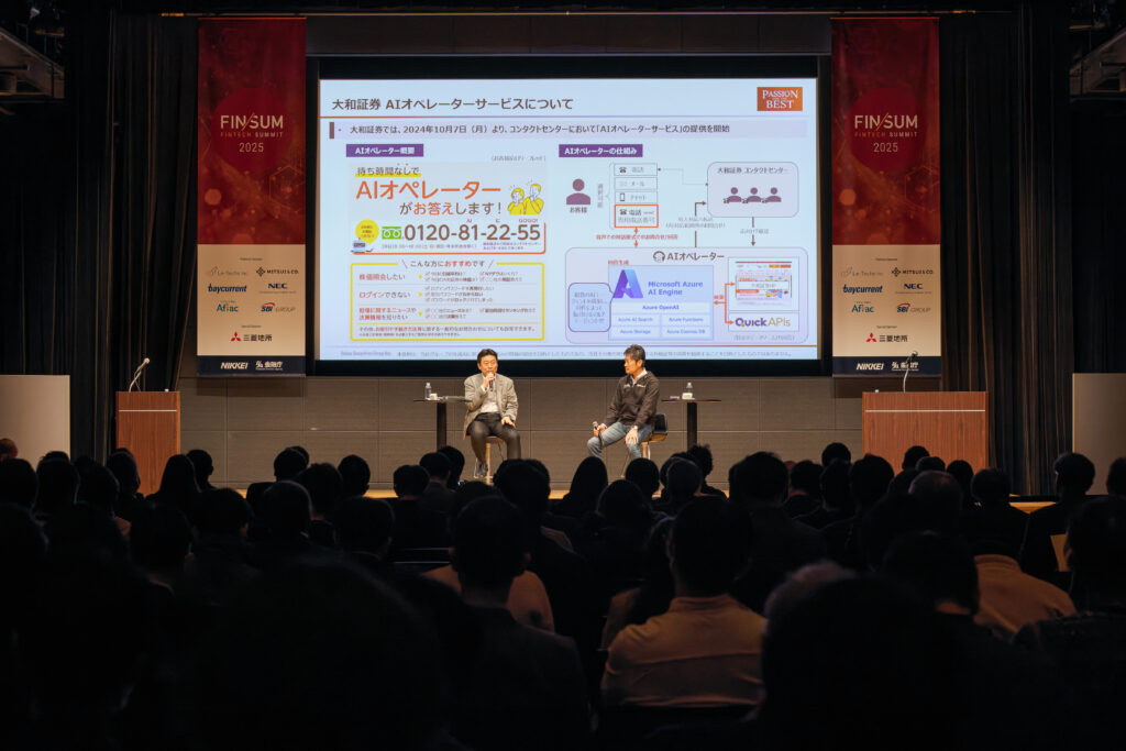 A man and woman sitting on stage in front of a large screen