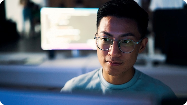 コンピュータモニター越しに見える、デスクで作業する人物。作業スペースは柔らかな光に包まれ、背景には大きなスクリーンが配置されている。