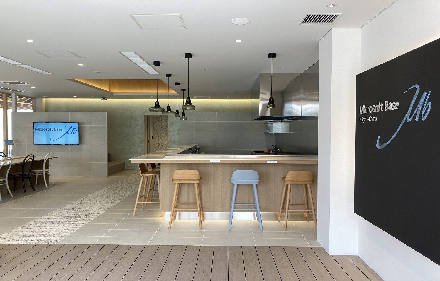 ダークな壁に「Microsoft Base Niigata-Kamo」と表示されたモダンなオープンラウンジスペースで、キッチンカウンター、木製バースツール、ペンダントライト、Microsoft Baseのロゴを映すテレビが設置されています。