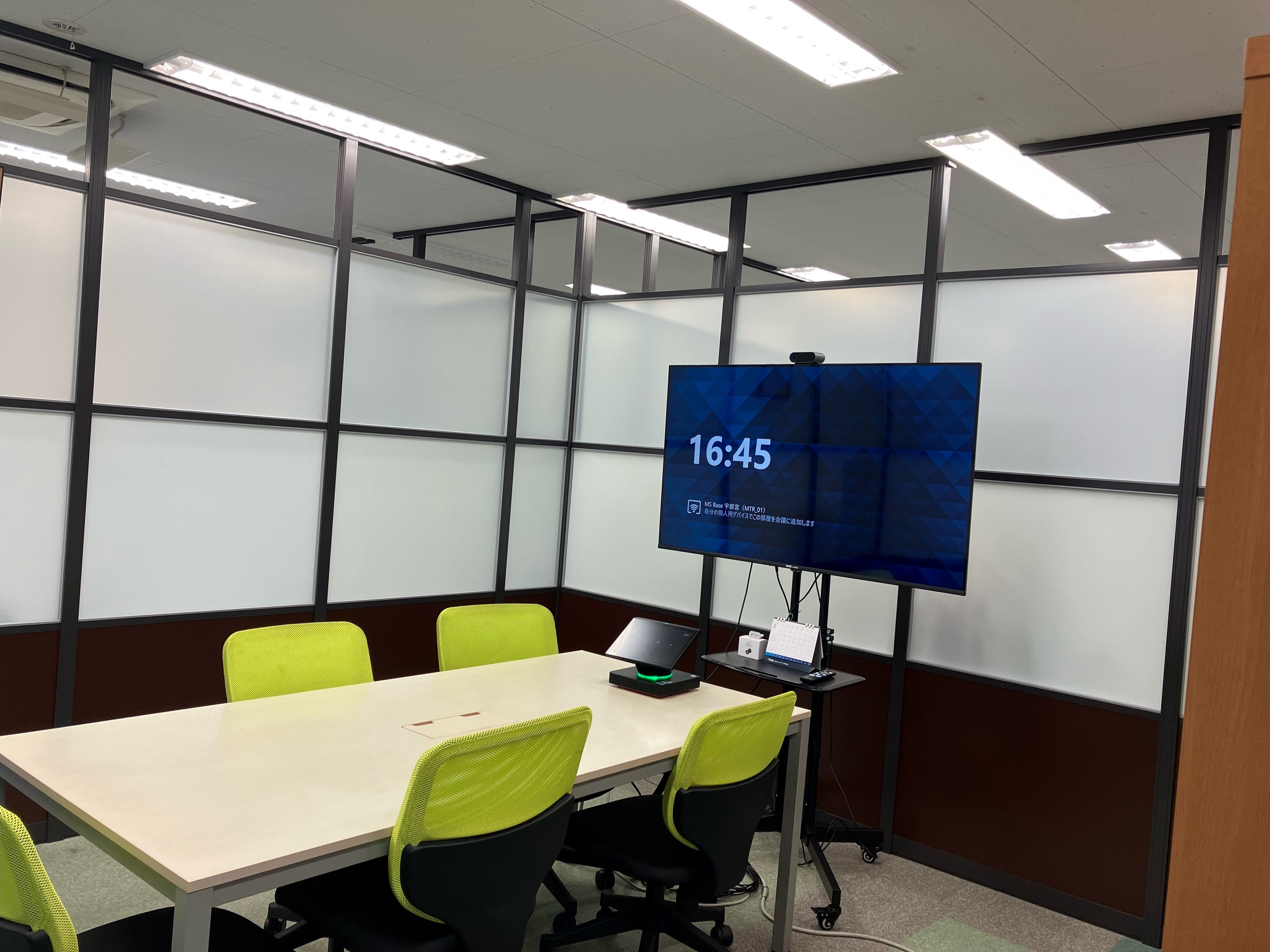 ガラス張りの小さな会議室。テーブル、ライムグリーンの椅子、スタンド付きの大型モニターが設置されている。