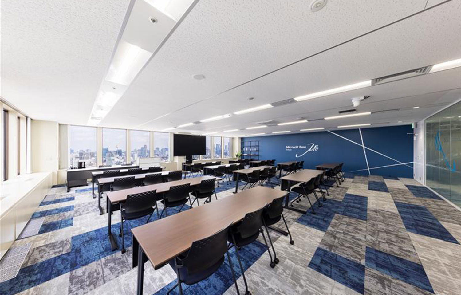 テーブルと椅子が並ぶ広々としたセミナールーム。青い模様のカーペット、大きな窓からの都市の眺望、そして「Microsoft Base」のロゴがある青いアクセントウォールが特徴。
