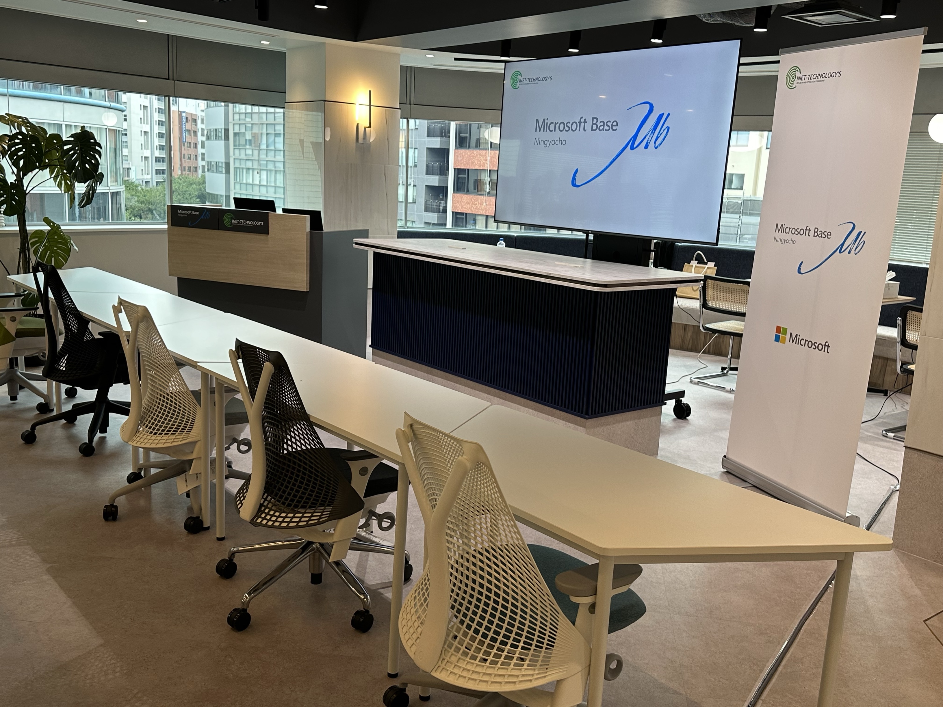 白いデスクとメッシュチェアが並ぶモダンな研修室。受付カウンターと「Microsoft Base」と表示された大型スクリーン、Microsoftブランドのスタンディングバナーが設置され、広い窓からは都市の建物が見える | INET Technology