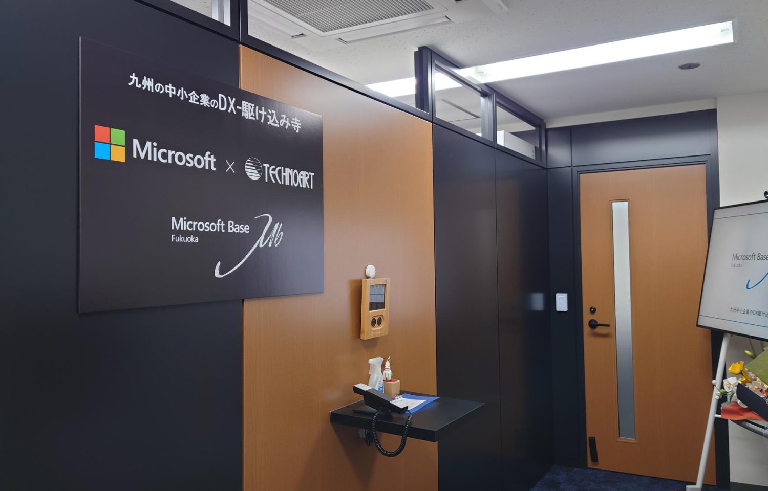 「九州の中小企業のDX-駆け込み寺」と書かれた看板があり、「Microsoft × TECHNOART」および「Microsoft Base Fukuoka」のブランドがダークな壁に表示されたオフィスのエントランスです。