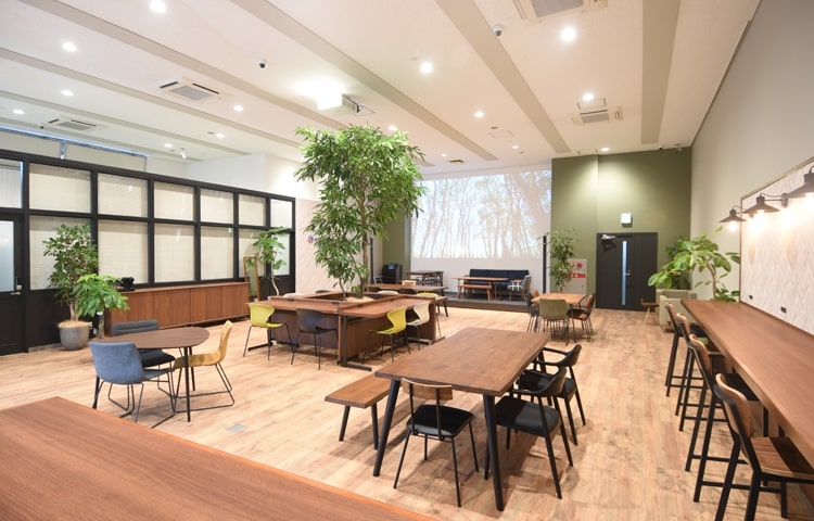 木製テーブルと多様な座席、大きな室内樹木、奥に大型投影スクリーンがある広々としたコワーキングラウンジ。