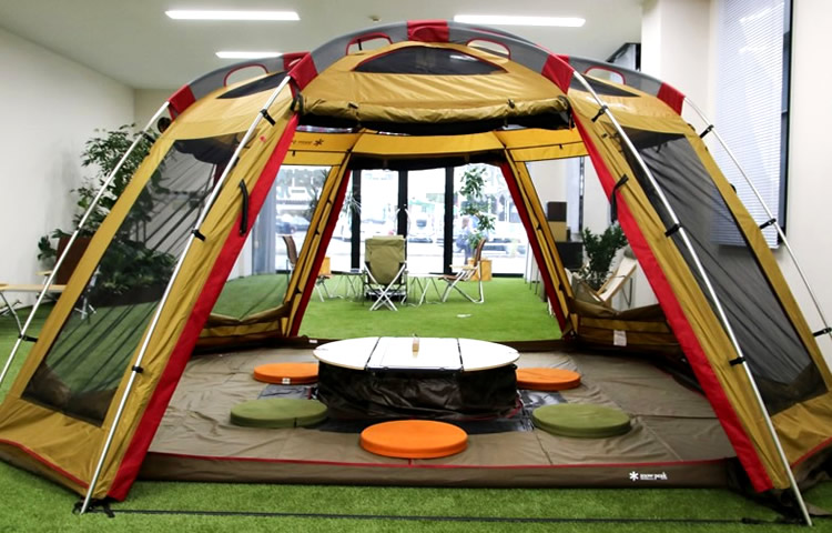 大きなドーム型テントと低めの円形テーブル、カラフルな座布団が人工芝に置かれた室内キャンプ風ラウンジ。
