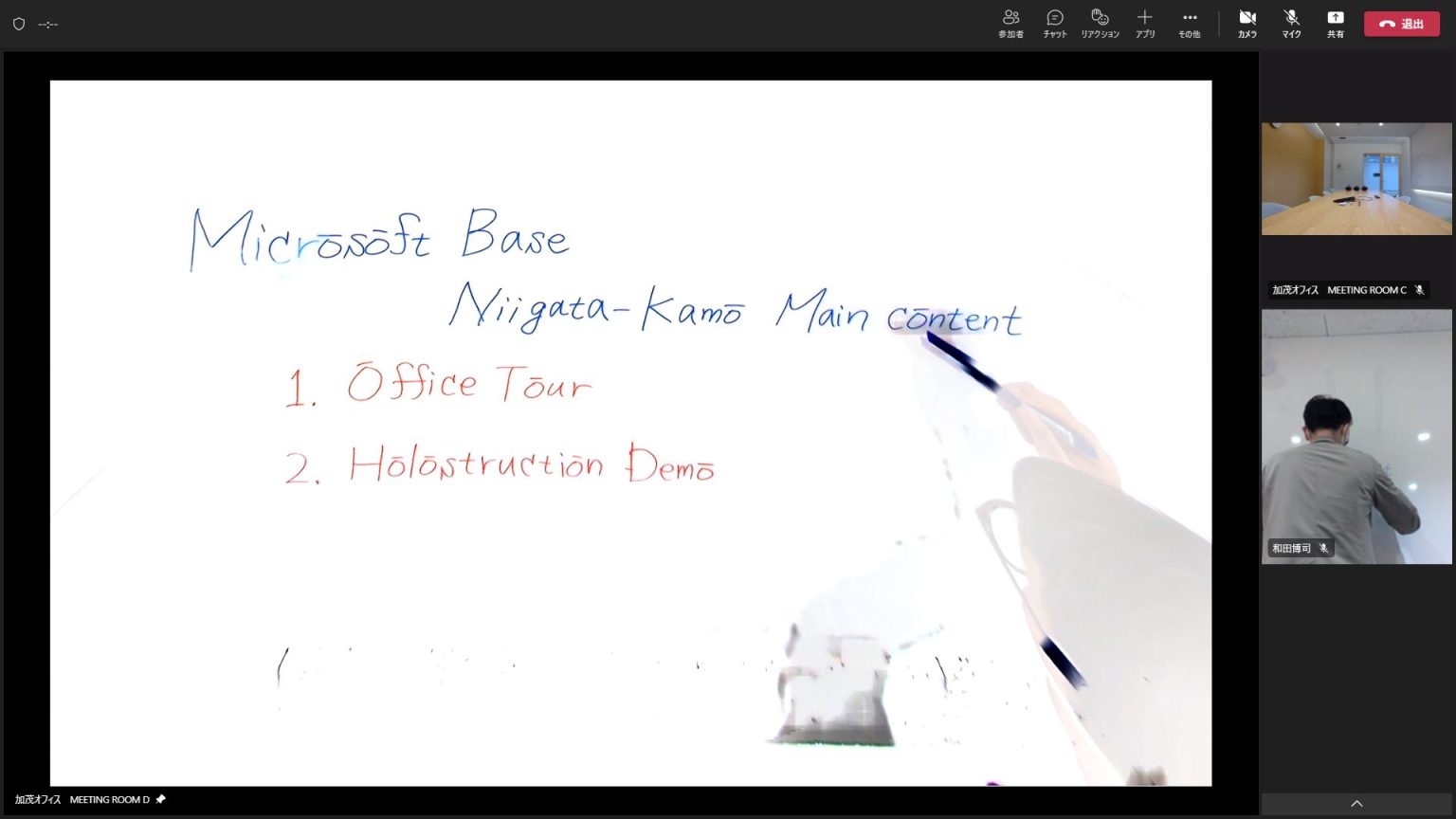 ビデオ通話中のデジタルホワイトボードプレゼンテーションのスクリーンキャプチャ。メインのホワイトボードには青と赤の手書き文字で「Microsoft Base」「新潟加茂 メインコンテンツ」「1. オフィスツアー」「2. ホロストラクションデモ」と記載されている。手でスタイラスを持っており、「メインコンテンツ」の文字に操作している様子が見える。右側には二つの小さなビデオ映像があり、参加者が表示されている。上の映像には「加茂オフィス MEETING ROOM C」とラベル付けされた空の会議室が映り、下の映像には「加茂オフィス MEETING ROOM D」とラベル付けされた白い壁の前に立つ人物の後ろ姿が映っている。インターフェースの上部バーにはさまざまな操作アイコンと日本語のテキストが含まれている。