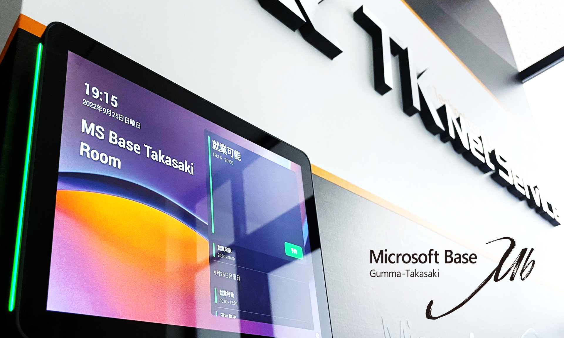 部屋の予約状況を示す壁面のデジタル表示パネルと、近くに「マイクロソフト ベース群馬高崎、19:15 MS ベース高崎ルーム」と書かれた看板 | 19:15 2022年9月25日日曜日。