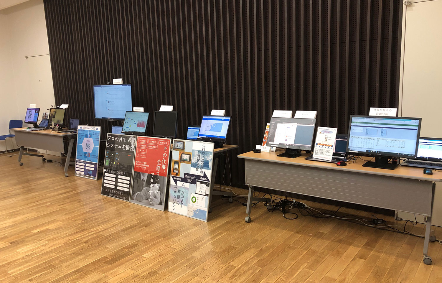 Microsoft 365、セキュリティ、モバイル、データ分析ソリューションを紹介する複数のモニターと展示パネルが並ぶ、展示会形式のテクノロジー紹介エリア。