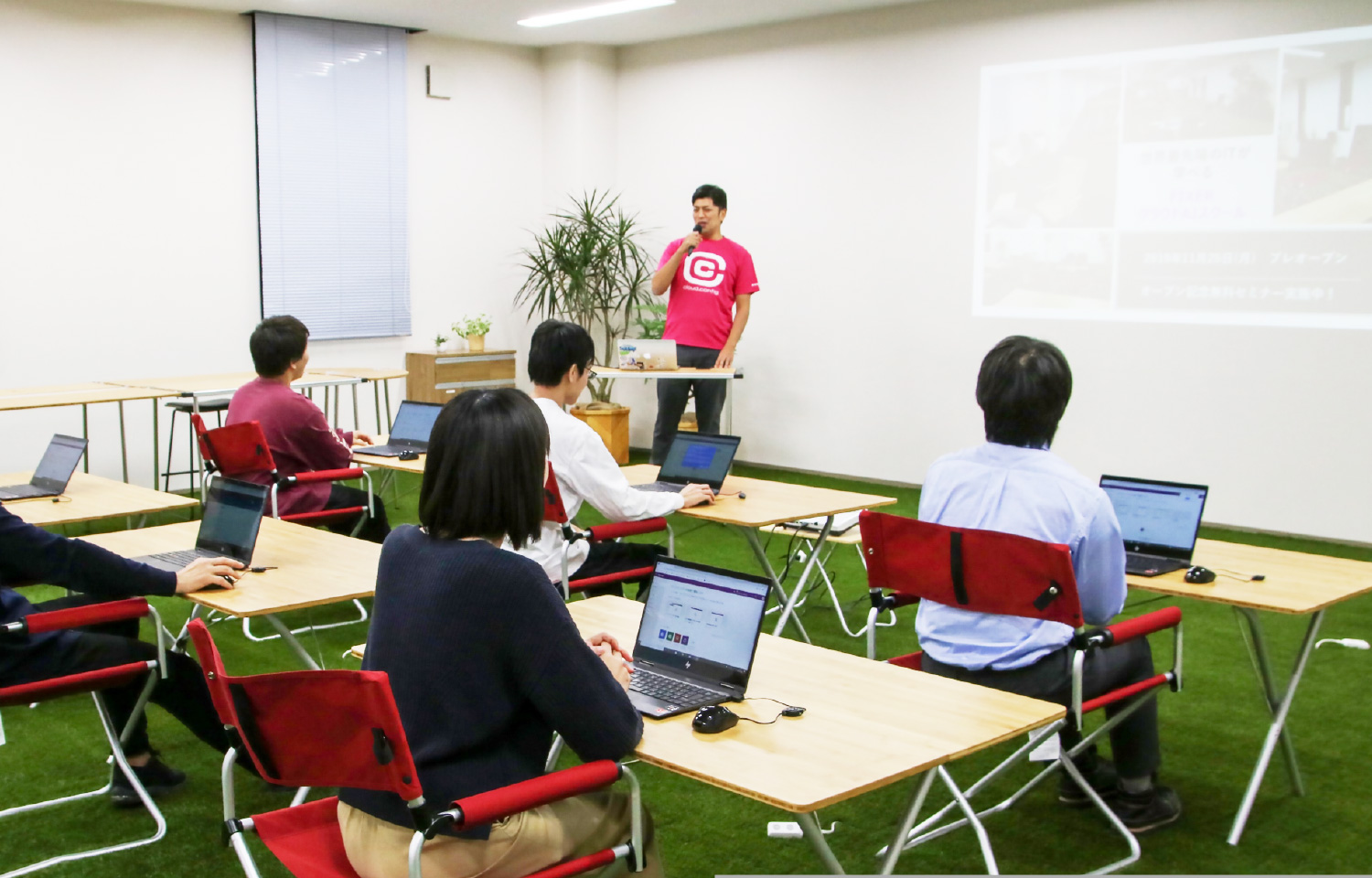 テーブルが並び参加者がノートPCを使う小規模な研修室、前方で講師がプレゼンテーション中。
