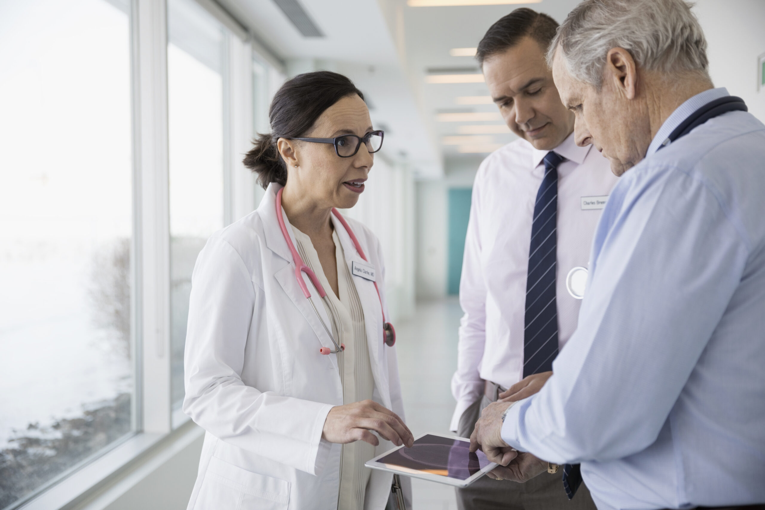 Doctor consulting with two professionals in a bright hospital corridor using a tablet. Health care; Microsoft Cloud for Healthcare; health solutions; clinical applications; physician; collaboration; teamwork. Usage approved only for Cloud for Healthcare applications