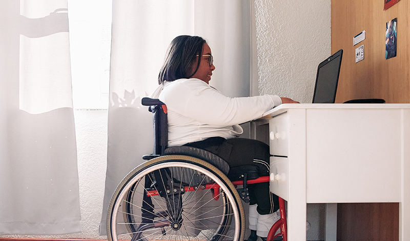Uma mulher em uma cadeira de rodas utiliza um notebook sobre uma mesa